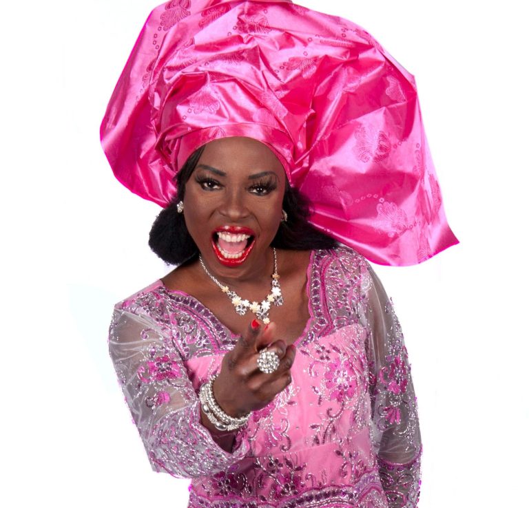 Drag queen in pink gown and headress smiling and pointing outwards